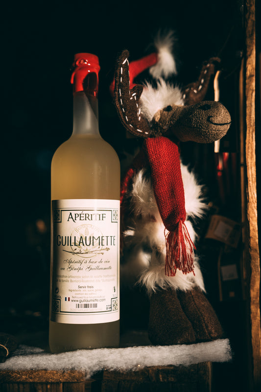 Bouteille de Guillaumette, apéritif au génépi, mise en scène dans une ambiance hivernale chaleureuse avec un renne en peluche vêtu d’un bonnet et d’une écharpe rouges, posée sur un lit de fausse neige.