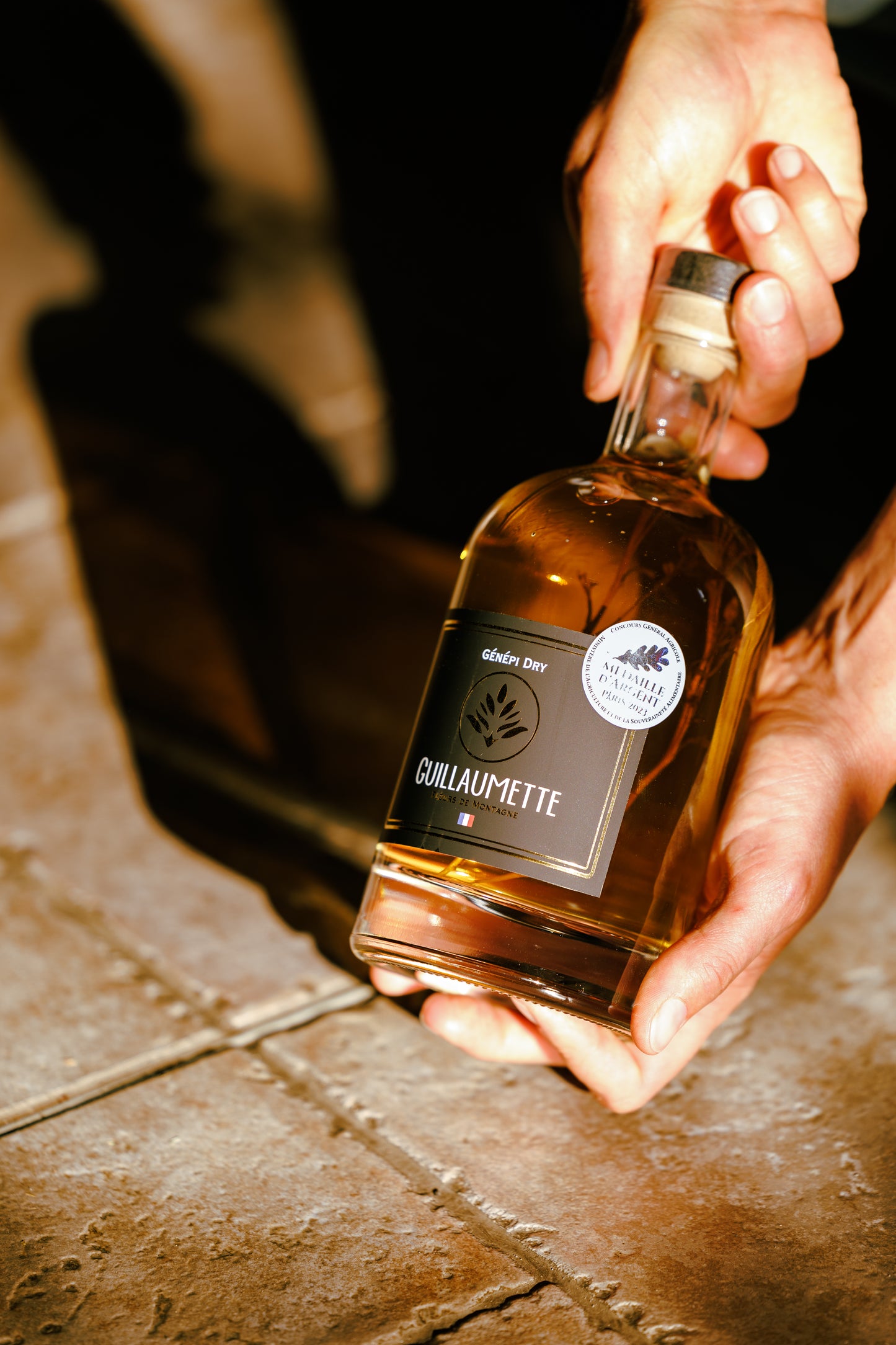 Bouteille de 70cl de Génépi Dry Guillaumette posée au sol et tenue en main, spiritueux de montagne artisanal médaillé d’argent.