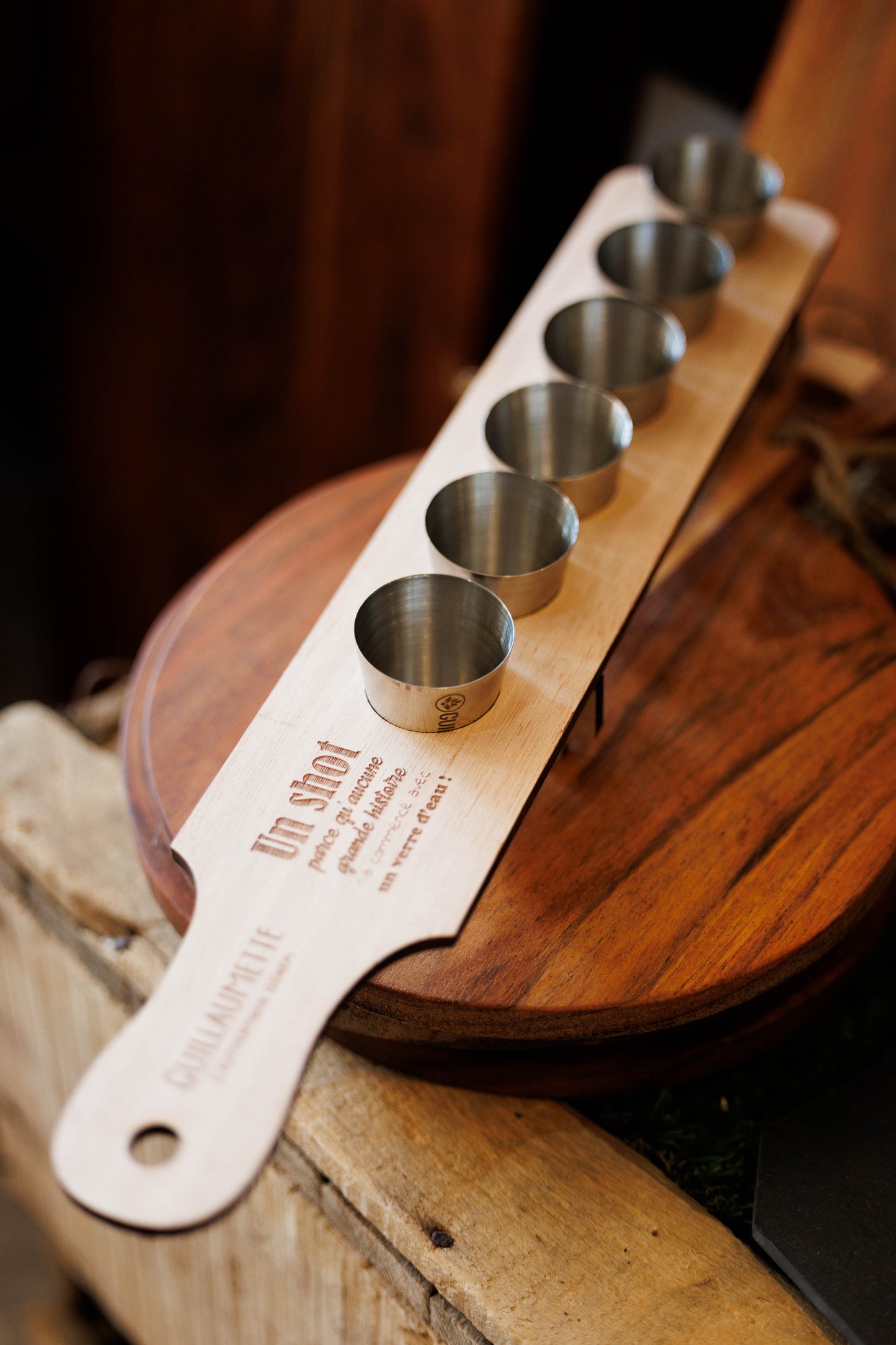 Planche à shot en bois gravée d’une citation humoristique, avec 6 verres à shot en inox Guillaumette posée sur un présentoir en bois.