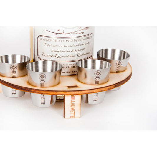 Set de verres en inox Guillaumette disposés dans un support circulaire en bois, posé autour d’une bouteille de génépi artisanal Guillaumette. Le support et les verres sont gravés avec soin pour refléter l’authenticité et la tradition du produit.