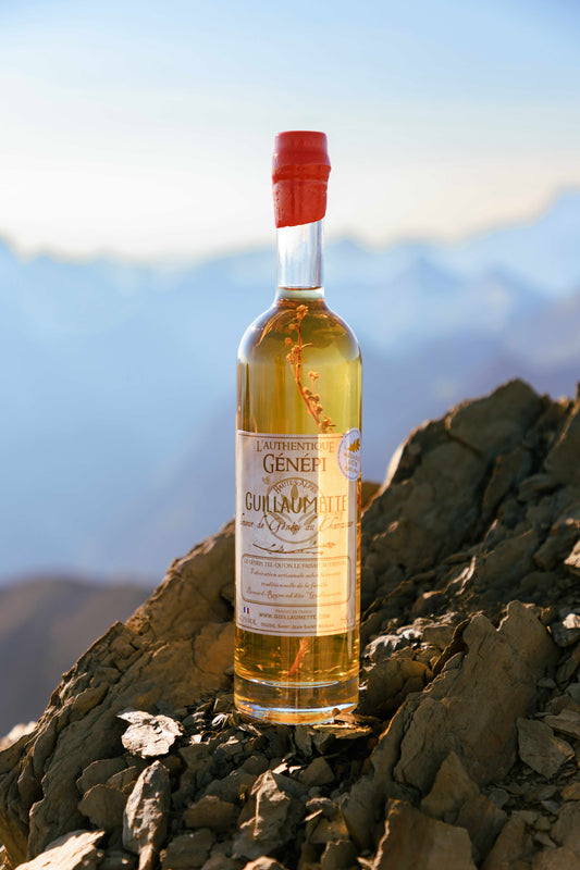 Bouteille de Génépi Guillaumette posée sur un rocher en haute montagne, avec vue sur les sommets au coucher du soleil.
