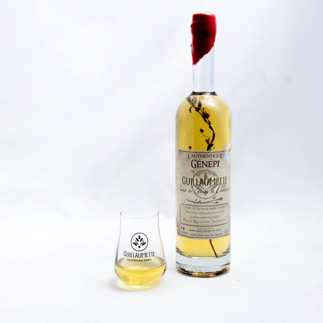 Bouteille de Génépi Guillaumette Authentique 70cl avec brin de plante visible et cire rouge sur le bouchon, accompagnée d’un verre à liqueur logotypé, rempli de Génépi, sur fond blanc