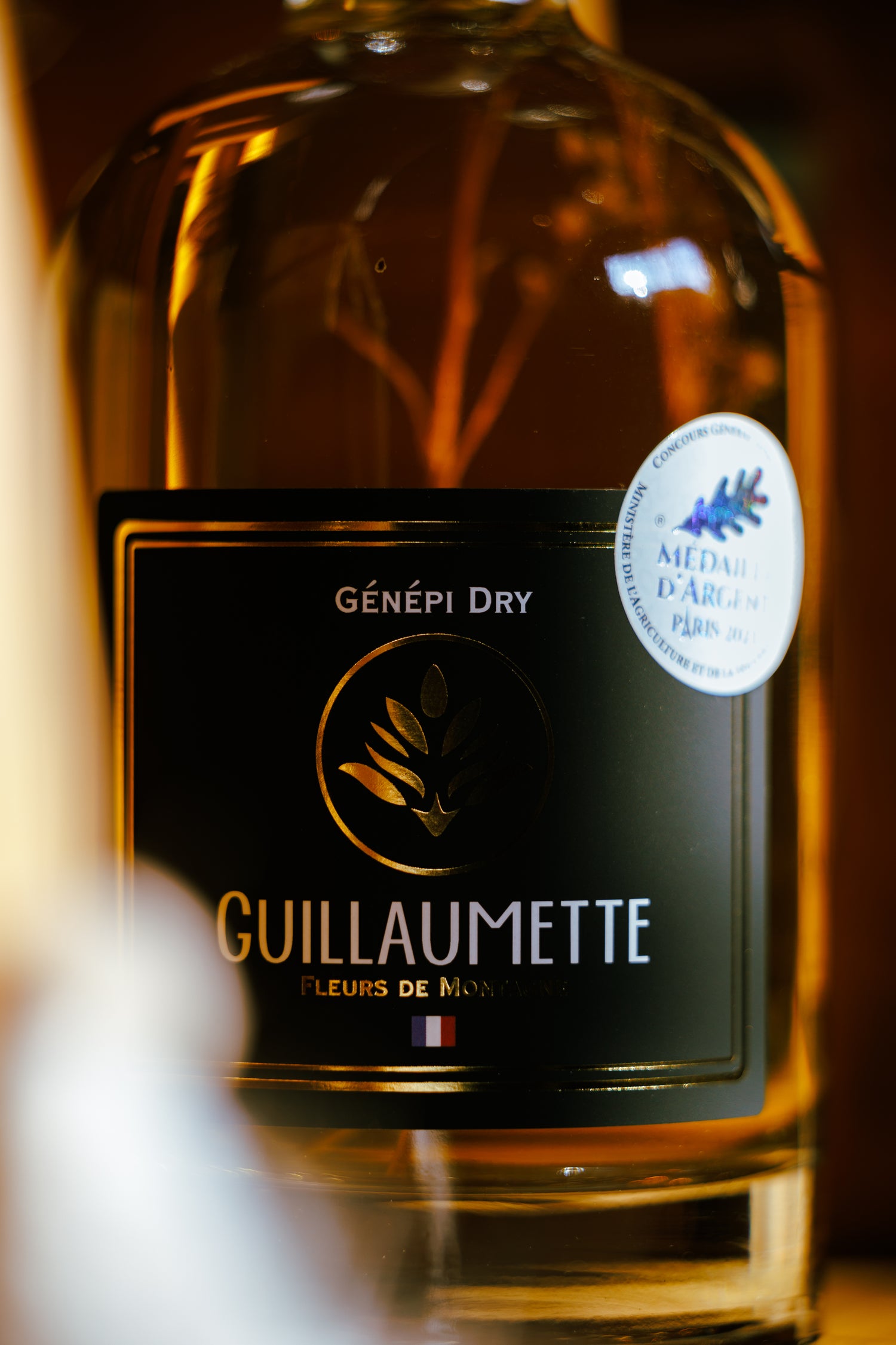 Zoom sur l’étiquette de la bouteille de Génépi Dry Guillaumette 70cl, spiritueux artisanal médaillé d’argent, avec détails dorés et fleurs de montagne dans la bouteille.