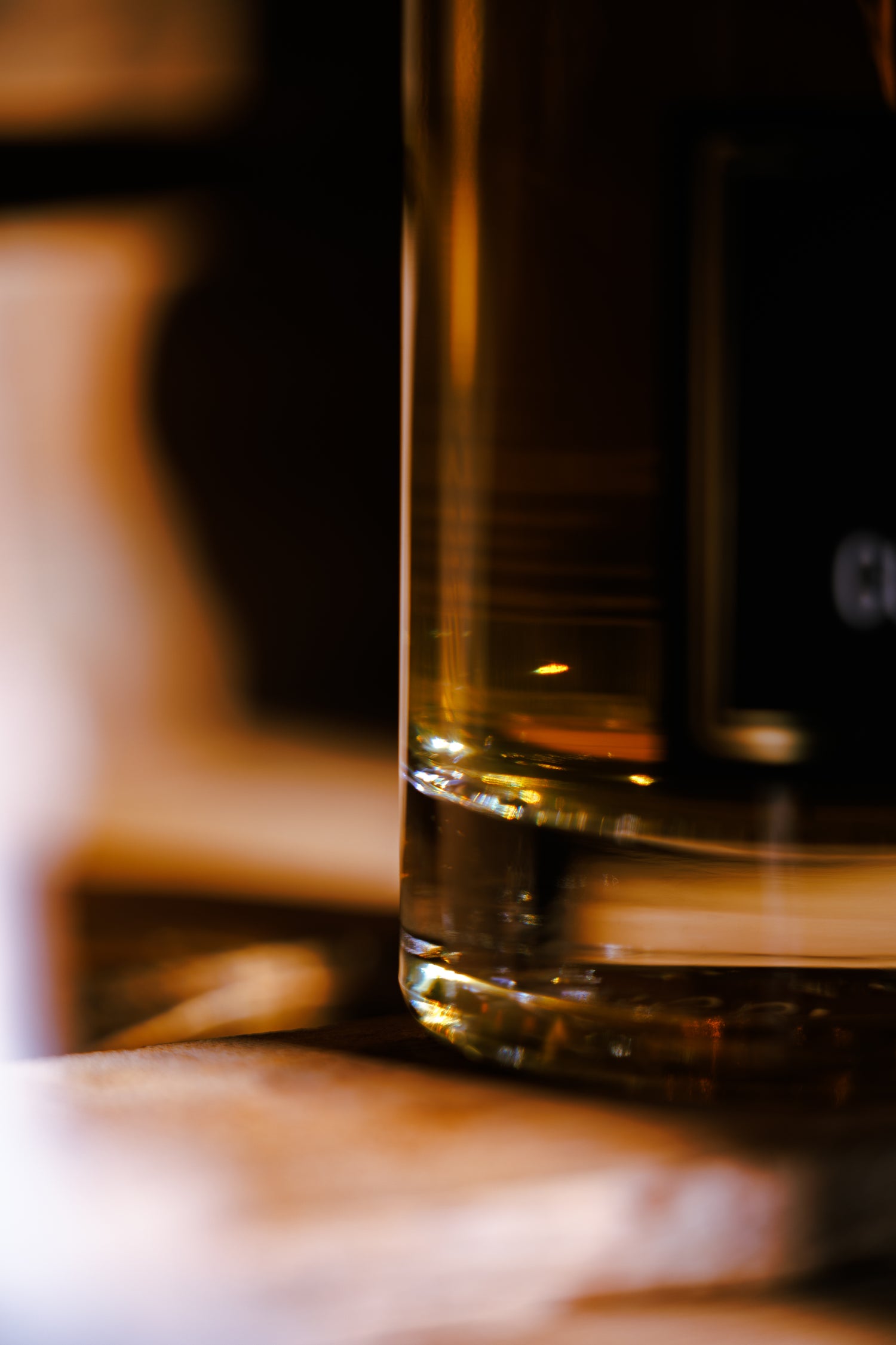 Gros plan sur la base d’une bouteille de Génépi Dry Guillaumette 70cl posée sur une table en bois, avec des reflets dorés chaleureux